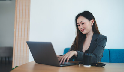 Asian young beautiful happy professional successful businesswoman designer sitting smiling at workstation desk using laptop notebook computer and smartphone working remotely online at home office