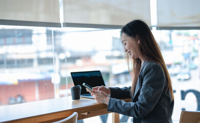 Fototapeta premium Asian young beautiful happy professional successful businesswoman designer sitting smiling at workstation desk using laptop notebook computer and smartphone working remotely online at home office