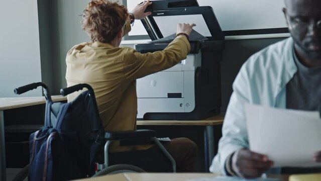 Young man in wheelchair using printer to copy business papers while working in office