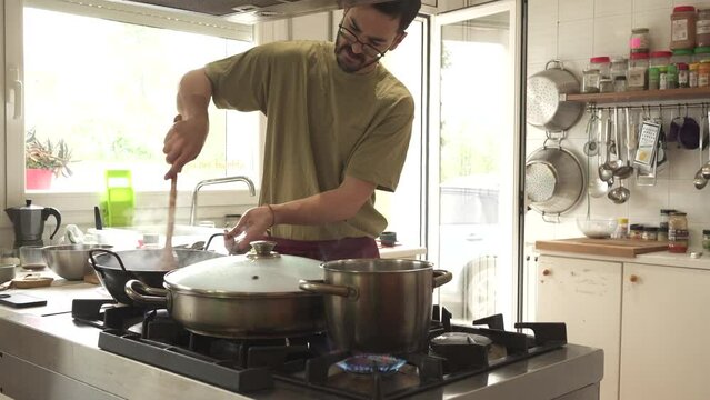 man cooking with several large pots, frying pan and boiling pot on gas fire. dressed in maroon apron on skirt and green T-shirt.
