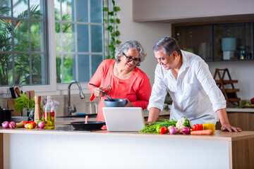 Indian asian senior old age cheerful couple using tablet computer or laptop in the kitchen while cooking