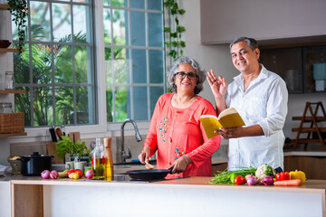 Senior Indian asian couple cooking together husband reading a recipe from culinary book.
