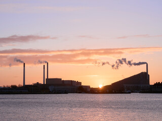Industriegebäude und rauchende Fabrikschlote am Wasser vor aufgehender Sonne und Morgenrot
