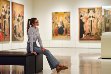 Thoughtful young Caucasian woman wearing glasses and looking at exhibition. Concept of Museum Day.