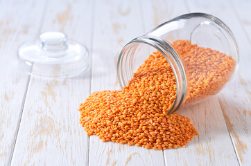 Raw red lentils spill out of a glass storage jar on a white wooden table.