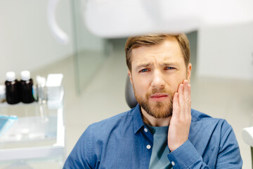 Sad man sitting in dental chair suffering from acute tooth pain, rubbing his cheek, close up