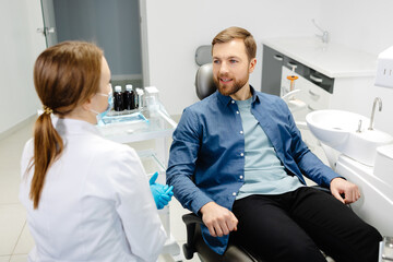 Blonde female dentist in dental office talking with male patient and preparing for treatment. Handsome bearded man in dentist chair looking at his doctor with smile