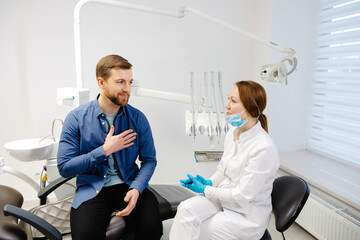 Obraz premium Blonde female dentist in dental office talking with male patient and preparing for treatment. Handsome bearded man in dentist chair looking at his doctor with smile