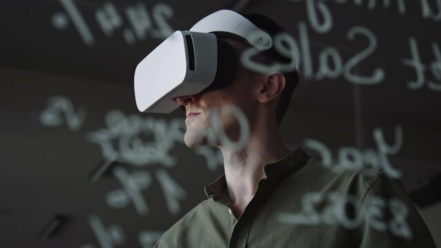 View through glass wall with written math formulas of man putting on VR headset and using remote controller while testing new app in office of big tech company