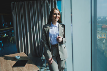 A business woman in a classic suit and a white shirt is standing by the window in a modern office. Modern business lady at the desk in the office