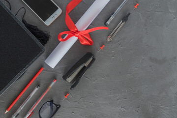 A black graduation cap, a rolled-up diploma with a red ribbon, pencils, pens, a stapler, a phone on...