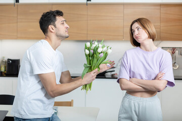 Young guilty husband kneeling and begging for forgiveness with a bouquet of flowers to his deadpan...