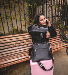 Stylish and young traveler latina brunette with fashion look and pink suitcase exploring city park while using the phone