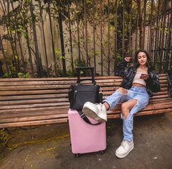 Stylish and young traveler latina brunette with fashion look and pink suitcase exploring city park while using the phone