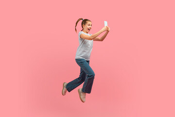 A young girl is captured mid-air, jumping energetically with a cell phone in hand. She appears...