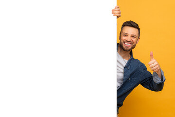 A man with a beard is standing and holding a sign in one hand while giving a thumbs up with the...