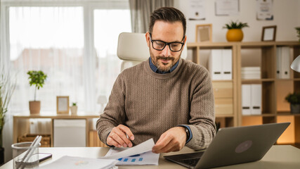 Adult businessman sit at office look on laptop hold paper at office