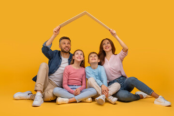 A smiling family sits closely together with the parents holding their arms up to form the shape of a house over their two children heads, all set against a vibrant yellow backdrop