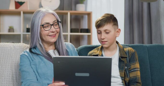 Pretty woman grandmother hugging her cute joyful grandson boy when they together using laptop computer at home