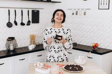 Cheerful woman standing in bright kitchen smiling laughing drinking hot beverage coffee tea, dressed in patterned pajama set. The kitchen counter arranged with ingredients, utensils, atmosphere 