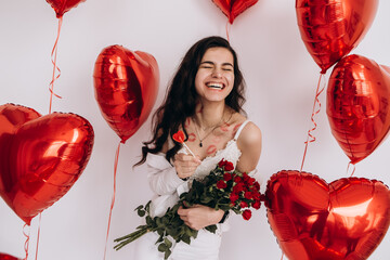 A smiling woman with kisses lipstick is holding a bouquet of red roses, candy, and standing amidst heart-shaped helium balloons, possibly celebrating a special occasion like Valentines Day
