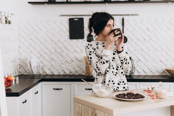 Cheerful woman standing in bright kitchen drinking hot beverage coffee tea, dressed in patterned pajama set. The kitchen counter arranged with ingredients, utensils, atmosphere domestic