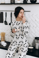 Cheerful woman standing in bright kitchen smiling eat candy, dressed in patterned pajama set. The kitchen counter arranged with ingredients, utensils

