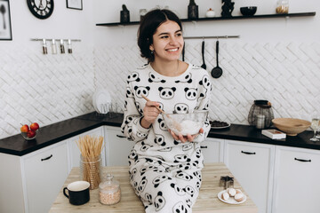 Cheerful woman sitting in bright kitchen mixing dough in a bowl, making homemade cookies, dressed in patterned pajama set. The kitchen counter arranged with ingredients, utensils, atmosphere domestic