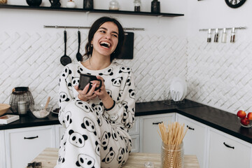 Cheerful woman standing in bright kitchen smiling drinking hot beverage coffee tea, dressed in patterned pajama set. The kitchen counter arranged with ingredients, utensils, atmosphere domestic
