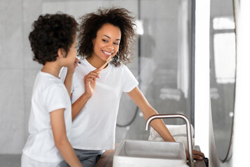 Black woman is seen standing next to a young boy, both brushing their teeth. Mother holds a...