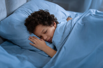 African American young boy is peacefully sleeping in a bed with blue sheets. He looks comfortable...