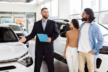 In a well-lit automobile showroom, a professional salesman, holding a blue clipboard, is warmly...