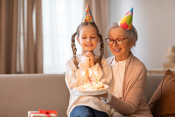 An older woman and a young girl sit on a couch, with a birthday cake in front of them. The woman is lighting the candles on the cake while the girl eagerly watches - Powered by Adobe