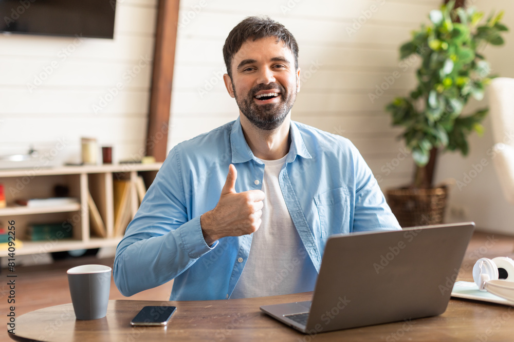Poster Handsome man is seated at a table, engaged with a laptop in front of him. He is expressing approval by giving a thumbs up gesture.