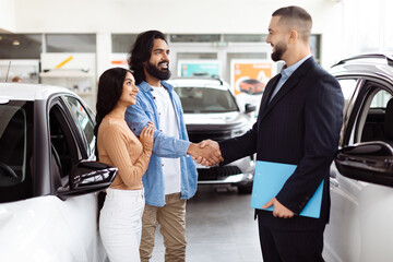 A young Indian couple is finalizing a deal with a car salesman inside a vehicle showroom. They seem pleased and are shaking hands with the agent, who is dressed in a business suit
