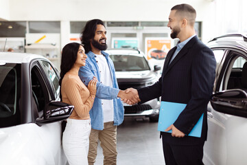 A young Indian couple is finalizing a deal with a car salesman inside a vehicle showroom. They seem...