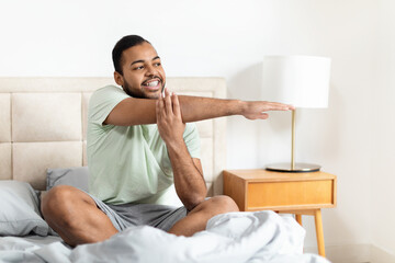 A young African American man is sitting on a bed, wearing casual sleepwear and stretching with a...