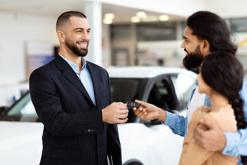 A well-dressed salesman is standing in a car showroom, presenting car keys to a cheerful Indian...