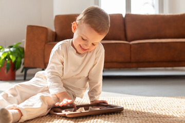 A young boy is sitting on the floor engrossed in playing on a tablet. He is focused on the screen,...