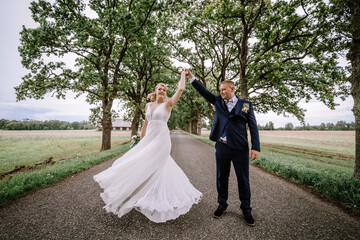 A bride in a flowing white dress and a groom in a blue suit are joyfully dancing on a tree-lined rural road. Their dance creates a dynamic, playful scene as they celebrate their wedding day...