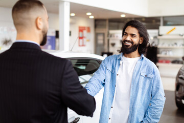 Two men are standing in a car showroom, shaking hands in agreement. The men appear serious and...