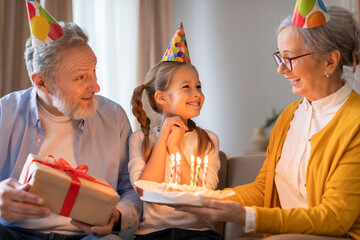 A young girl clad in a birthday hat smiles brightly as her grandparents, also donning festive party...
