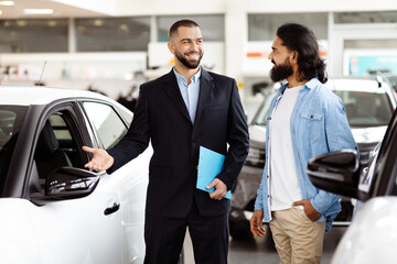 A smartly dressed car salesman, with a friendly demeanor, stands next to a shiny new car in a...