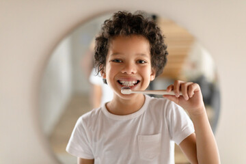 African American young boy is standing in front of a mirror, holding a toothbrush and brushing his...