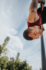 A fit man with visible tattoos engages deeply in a workout at an urban park, hanging from fitness equipment on a bright, sunny day.