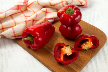 Cutting board with several whole and halves of red bell pepper and red kitchen towel on white wooden background. .