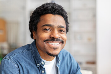 A cheerful young African American man with a mustache and curly hair is casually dressed in a denim...