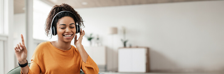 Great Sound. Smiling black woman touching wireless headset sitting on beanbag chair in living room,...