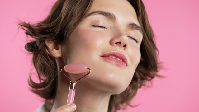 Young woman with rose quartz stone roller on pink background. Facial self care