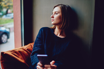 Thoughtful young woman dressed in casual wear looking out of window holding smartphone in...